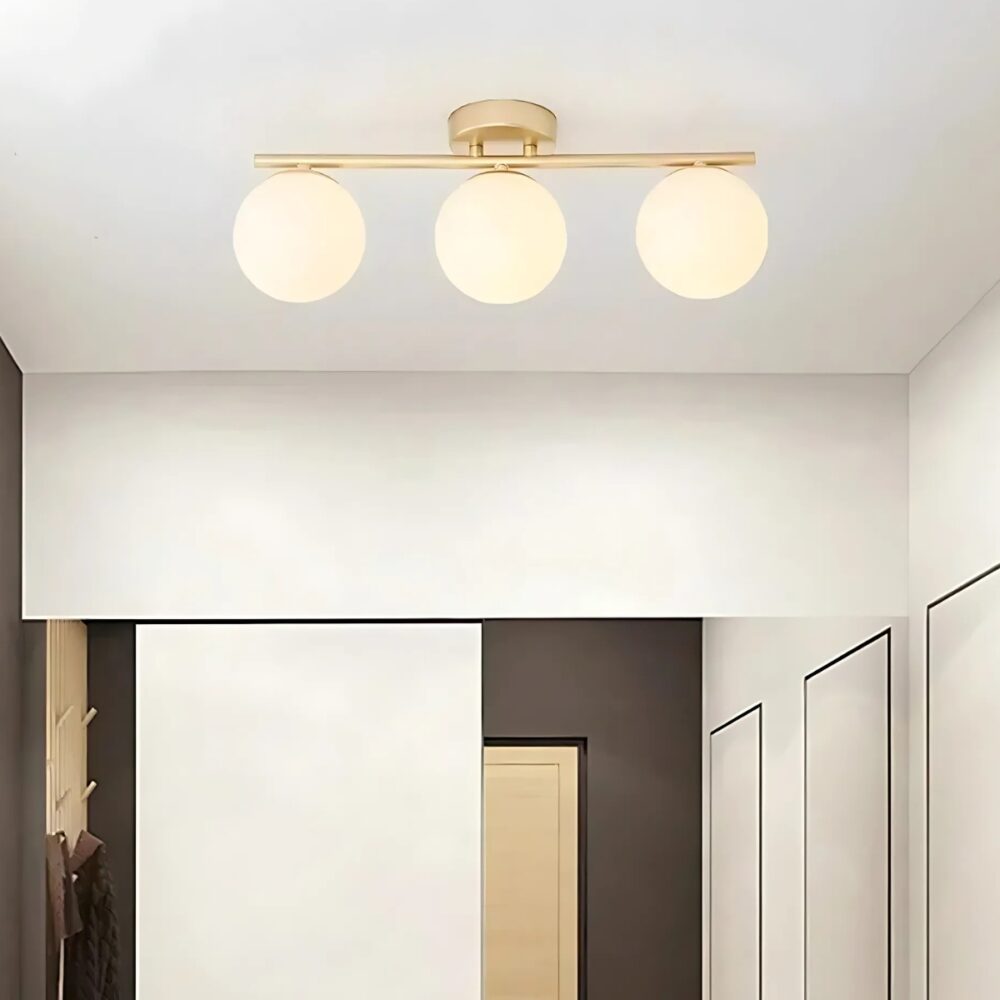 Lampe plafonnier avec boules lumineuses. Elle est dorée et composée de 3 boules fixées sur une barre horizontale. Fixée dans une pièce aux murs beiges.