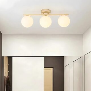 Lampe plafonnier avec boules lumineuses. Elle est dorée et composée de 3 boules fixées sur une barre horizontale. Fixée dans une pièce aux murs beiges.
