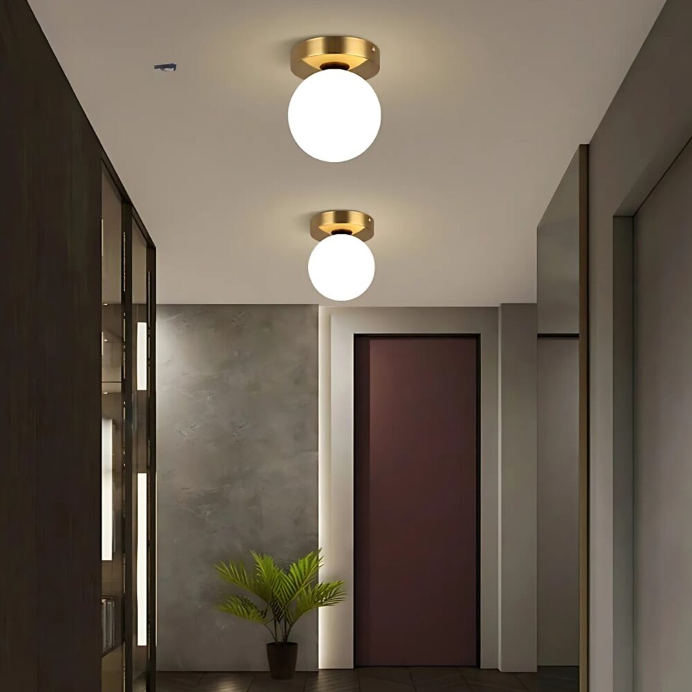 Lampe plafonnier boule lumineuse avec socle rond doré. Deux lampes l'une derrière l'autre fixées dans un couloir et allumées. Sur la gauche des plafonds marrons, en face une porte marron sur un mur gris et sur la droite un mur gris.