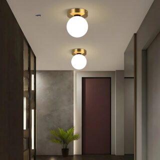 Lampe plafonnier boule lumineuse avec socle rond doré. Deux lampes l'une derrière l'autre fixées dans un couloir et allumées. Sur la gauche des plafonds marrons, en face une porte marron sur un mur gris et sur la droite un mur gris.