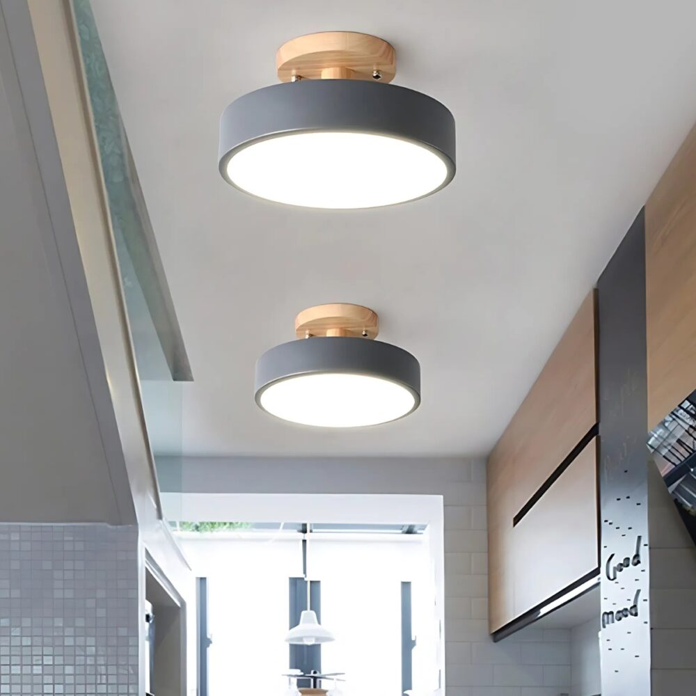 Lustre plafonnier plat au design nordique de couleur grise avec un support imitation bois fixé au plafond. L'éclairage blanc est diffusé par la partie en acrylique plate. Deux lustres plafonniers identiques fixés l'un derrière l'autre dans une cuisine avec des placards beige et gris sur la droite et une fenêtre au fond.