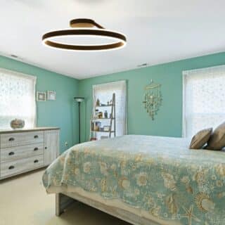Plafonnier chambre adulte cercle lumineux LED allumé et fixé dans une chambre aux murs verts. Sur la droite un lit deux places avec une literie dans les tons verts et motifs de coquillage. Sur la gauche, une commode à tiroirs.