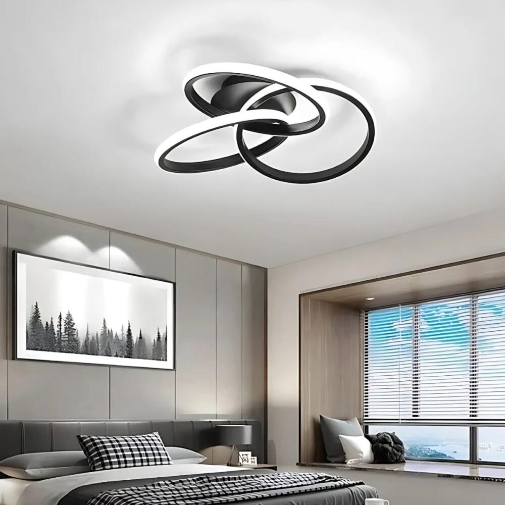 Plafonnier design anneaux entrelacés de couleur noire fixé dans une chambre d'adultes. Sur la droite une grande fenêtre, à gauche un lit double avec au-dessus un cadre noir et blanc.