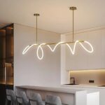 Plafonnier design avec tube souple lumineux enroulé sur la structure suspendue dorée. Il est fixé au-dessus d'un comptoir blanc où des fauteuils hauts gris sont installés. Au fond à droite, une cuisine.