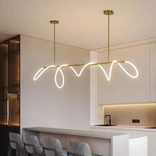 Plafonnier design avec tube souple lumineux enroulé sur la structure suspendue dorée. Il est fixé au-dessus d'un comptoir blanc où des fauteuils hauts gris sont installés. Au fond à droite, une cuisine.