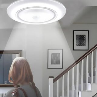 Plafonnier détecteur de mouvement à piles qui éclaire une dame qui passe devant un escalier avec les barreaux blancs et la rambarde en bois. Deux cadres fixés au mur dans l'escalier.