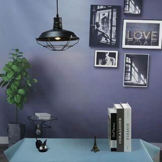 Plafonnier industriel suspendu en métal noir de forme ronde style cage avec une grille en-dessous. Fixé au-dessus d'une tble rectangulaire bleu avec des livres posés dessus. Le mur de derrière est bleu et des cadres y sont accrochés.