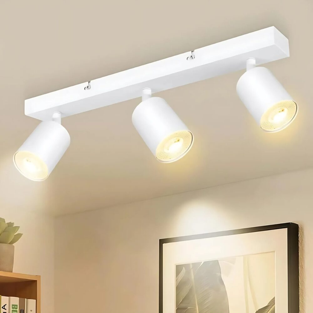 Plafonnier spots orientables blanc fixé sur un plafond beige. Les 3 spots sont munis d'une ampoule allumée. On aperçoit un tableau sur le mur de derrière.