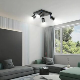 Plafonnier spots orientables noirs avec 4 têtes sur un support carré noir. Fixé dans un salon avec un canapé gris. Une fenêtre sur le pan de droite.