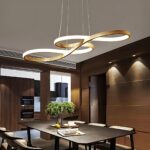 Plafonnier suspendu aux courbes fluides de couleur dorée avec la bande lumineuse sur l'ensemble des courbes. Il est fixé au-dessus d'une table rectangulaire en bois avec des chaises autour. Les murs de la pièces sont marron.
