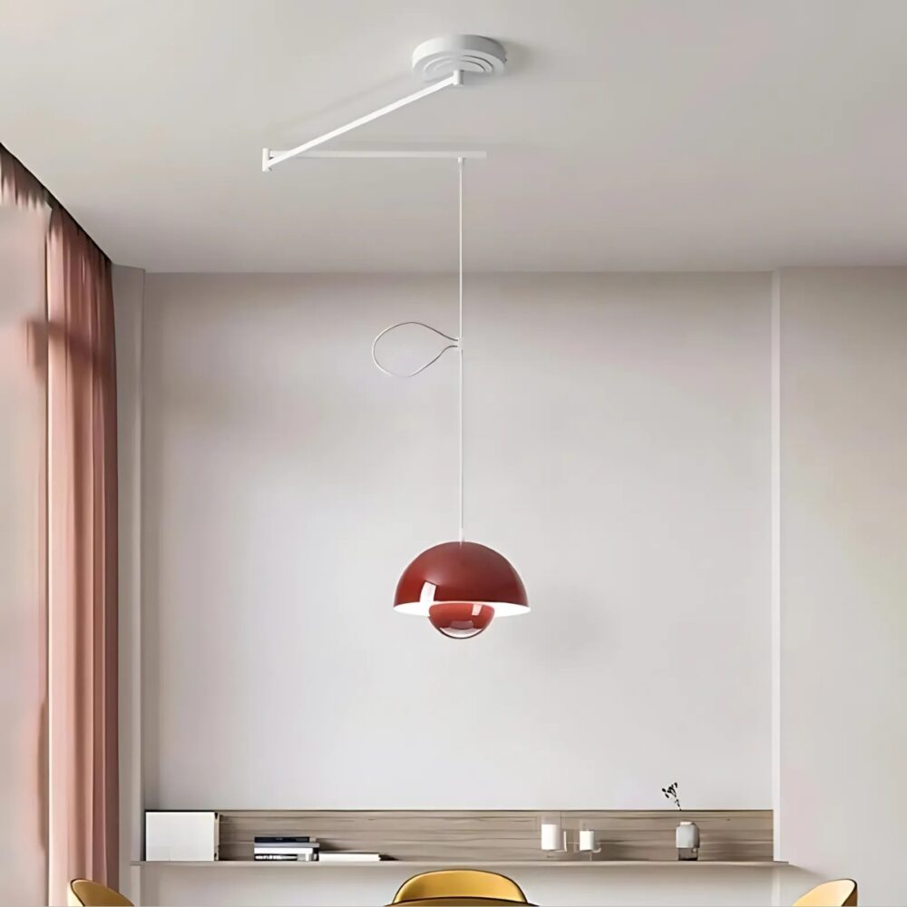 Plafonnier déporté moderne coloré avec un bras orientable en 2 parties. L'abat-jour rond est de couleur rouge avec la lumière reflété à l'intérieur. Il est suspendu par un câble blanc relié aux bras pliables. Dans une pièce aux murs blancs avec une baie vitrée sur la gauche, une table ronde et des sièges jaunes sous le plafonnier.