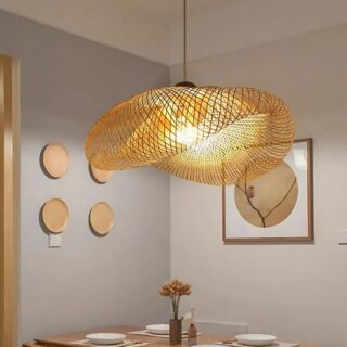 Plafonnier salle à manger en rotin naturel de forme ronde ressemblant à un chapeau recourbé vers le bas. Une ampoule au centre diffuse une lumièrer jaune. Suspendu dans une pièce avec un mur gris sur la gauche, un mur beige sur la droite avec un cadre. Une table se trouve sous le plafonnier.