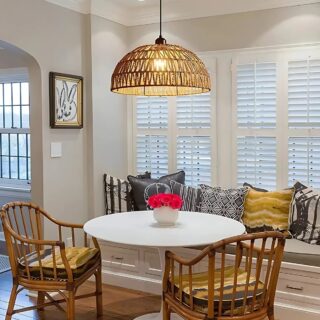 Lustre plafonnier en corde de chanvre de couleur beige naturel. L'ampoule au centre est allumée. Fixé au-dessus d'une table ronde blanche avec trois fauteuils en rotin autour de celle-ci. En fond, une fenêtre et un banc avec des coussins devant.
