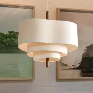 Lustre plafonnier en tissu style japonais de couleur beige avec 3 niveaux de tissu formant des cercles ondulés. Une tige couleur bois passe u centre reliée au câble de suspension. En fond, un mur avec deux tableaux de peinture.