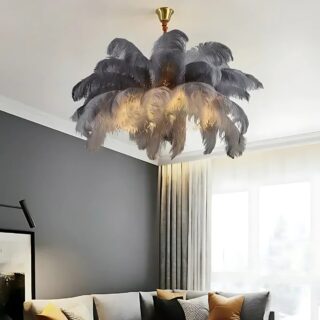 Plafonnier suspendu avec plumes colorées fixé dans un salon. Les plumes sont de couleur grise et la lumière jaune passe sous celles-ci. Sur la gauche, un mur gris avec un canapé et sur la droite une grande baie vitrée avec voilages blancs.