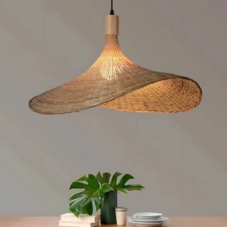 Plafonnier suspendu en forme de chapeau en rotin avec une lumière chaude diffusée par l'ampoule au centre. Sous le plafonnier, une table avec une plante et de la vaisselle blanche. Le mur au fond est gris.